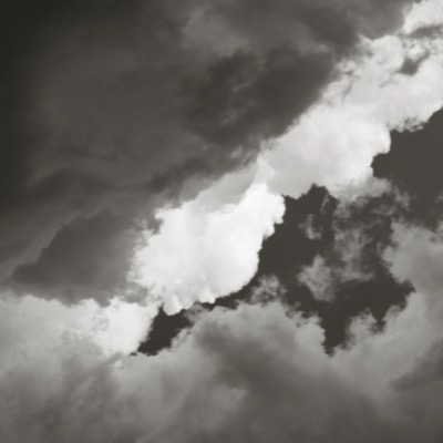 Black-and-white photograph of heavy storm clouds with a narrow break of bright cumulus light cutting diagonally across the frame.