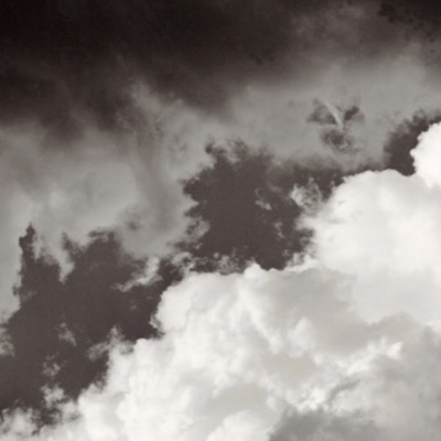 Black-and-white photograph of a grainy, high-contrast sky, dark storm clouds pierced by an intensely lit white cumulus tower in the lower right.
