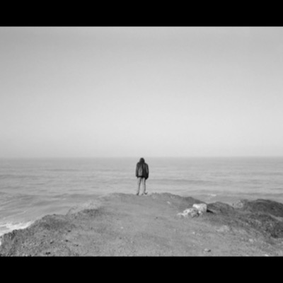Black-and-white photograph of a lone figure standing at the edge of a coastal bluff, seen from behind, looking out over a hazy ocean horizon with waves breaking on rocks below.