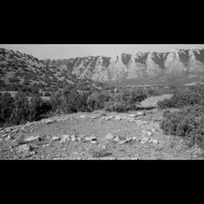 Black-and-white photograph of a tipi ring of weathered stones on a grassy, juniper-dotted slope in the Pryor Mountains, with layered sandstone cliffs rising behind.