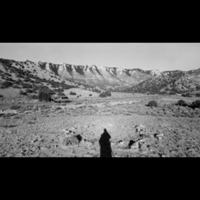 Black-and-white photograph of a parked minivan on a rural highway below sandstone cliffs, the photographer’s own shadow cast across a stone-scattered foreground.