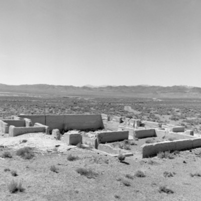 Black-and-white photograph of the crumbling concrete foundation walls of an abandoned building in open high desert, distant mountain ranges on the horizon.