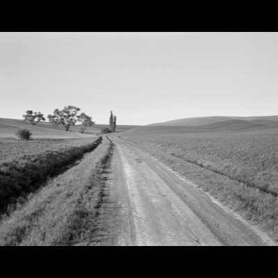 Black-and-white photograph of dirt tire tracks curving in tight rings around a fenced-off well pad in flat farmland, hills rising in the distance.