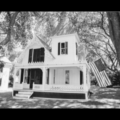 Black-and-white photograph of an ornate white Victorian gingerbread cottage with a turret and wraparound porch, a large American flag hanging from a shade tree beside it.