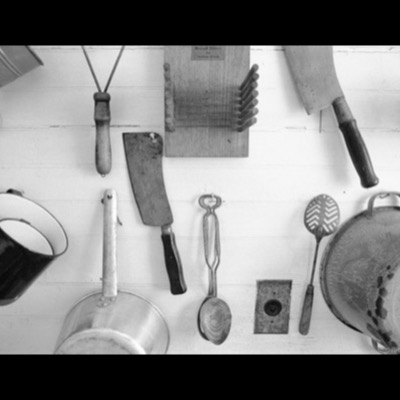 Black-and-white photograph of old kitchen tools hung on a white clapboard wall: cleavers, a bread slicer, tin cups, a saucepan, ice tongs, a slotted spoon, and a colander.