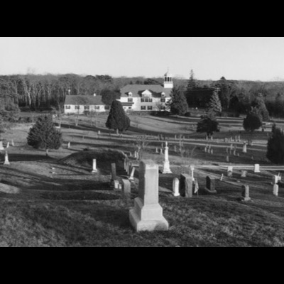 Black-and-white photograph of a hillside cemetery, rows of old gravestones descending toward a white cupola-topped town hall in the distance.