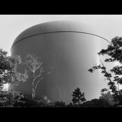 Black-and-white photograph of a massive domed industrial storage tank looming behind silhouetted trees, its curved metal surface filling most of the frame against a pale sky.