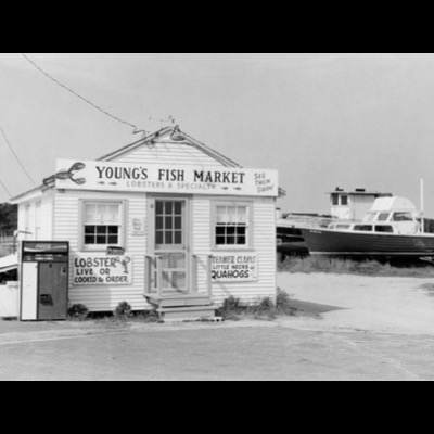 Black-and-white photograph of "Young’s Fish Market," a hand-painted roadside seafood shack advertising lobster, clams, and quahogs, a Coca-Cola machine out front and a boat parked in the lot behind it.