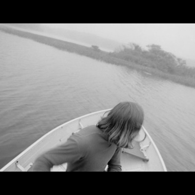 Black-and-white photograph, from behind, of a person leaning over the bow of a small boat crossing a calm inlet toward a marshy, tree-lined shore.