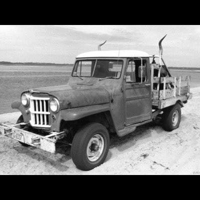 Black-and-white photograph of a weathered vintage Willys stake-bed truck with curved horns mounted on its roof, parked on a sandy beach beside calm water.