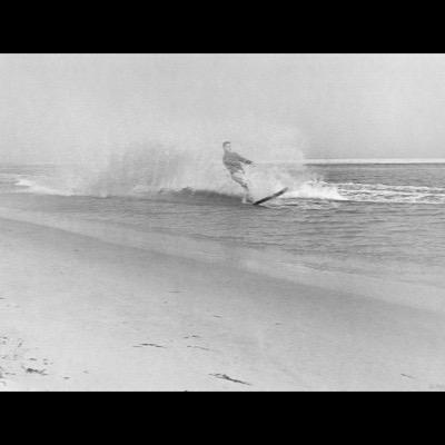 Black-and-white photograph of a skimboarder throwing up a spray of water as he cuts across the wash of a breaking wave near shore.