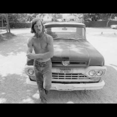 Black-and-white photograph of a shirtless, bearded man with a cigarette leaning against a vintage Ford pickup with a "TOURISTS GO HOME!" bumper sticker on the grille.