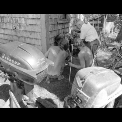 Black-and-white photograph of two shirtless young men and an older man crouched together working on equipment beside a weathered shed, vintage Johnson and Evinrude outboard motors in the foreground.