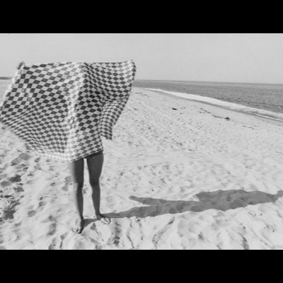 Black-and-white photograph of a person on a beach shaking out a large checkered blanket that billows over their head in the wind, ocean surf visible along the shoreline behind them.