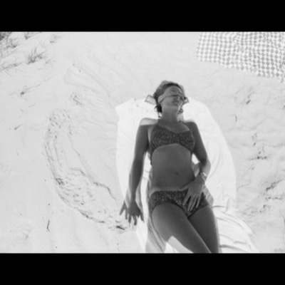 Black-and-white photograph, from directly above, of a woman in a bikini sunbathing on a dune, a scarf tied over her eyes and a checkered blanket in the corner of the frame.