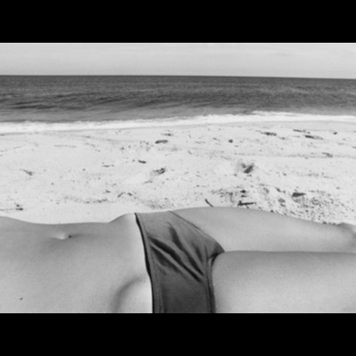 Black-and-white photograph, from behind, of a person’s bikini-clad lower back lying on the beach, filling the foreground below a view of the shoreline and gentle surf.