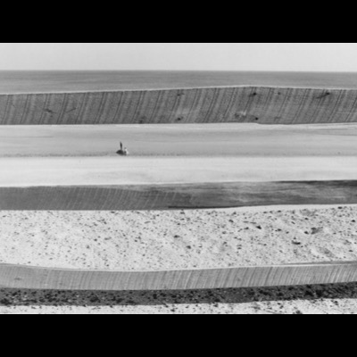 Black-and-white photograph, framed through parallel wooden groins stepping down a beach, of two tiny figures standing far below on a wide expanse of sand facing the sea.
