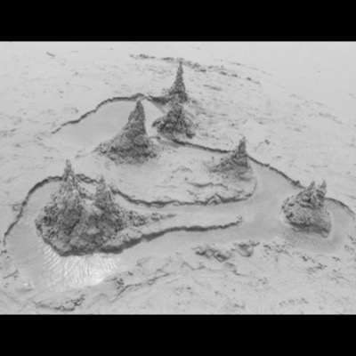 Black-and-white photograph, from above, of drip-cast sand spires shaped like miniature mountains, encircled by a winding channel of shallow water on a beach.