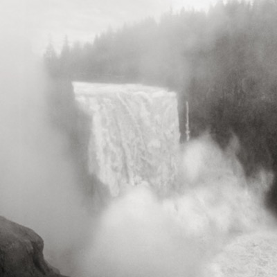 Black-and-white photograph of a broad dam spillway feeding a churning river, four visitors watching from a railed overlook as a highway bridge crosses in the background.