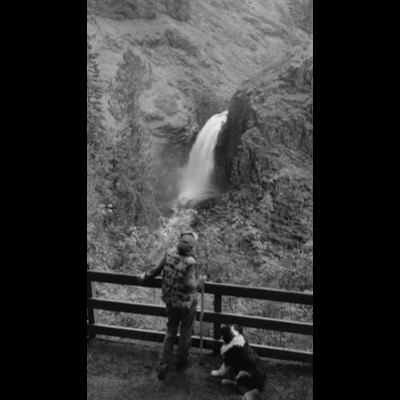 Black-and-white photograph of a small waterfall dropping through a dense conifer forest below fog-shrouded mountain ridges.