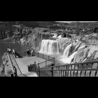 Black-and-white photograph, obscured by heavy waterfall spray, of a canyon and cultivated valley floor lined with poplar trees below layered desert cliffs.