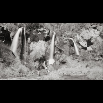 Black-and-white photograph, from below, of five swimmers or bathers on rocks beside a waterfall, one lying with legs raised near the plunge pool, sunlight streaking through the forest canopy above.