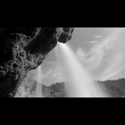 Black-and-white photograph looking straight up through overhanging rock into bright, blown-out waterfall mist against the sky.