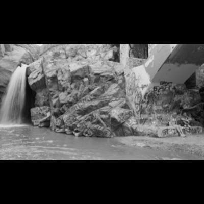 Black-and-white photograph of a rock cairn stacked at the edge of a forest clearing beside a narrow waterfall streaming down a distant cliff.