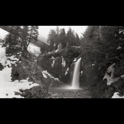 Black-and-white photograph, taken at an angle, of a waterfall pouring from a rocky ledge into a parking-lot overlook and highway visible in the valley below.