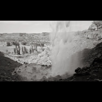 Black-and-white photograph of a broad waterfall descending in braided channels over layered dark rock into a still pool.