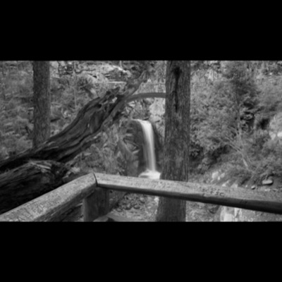 Black-and-white photograph, taken sideways, of a wide waterfall spilling down a rock face beside a wooded parking area and winding mountain highway below.