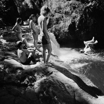 Black-and-white photograph of eight young people bathing and diving nude at the base of a sunlit forest waterfall, some standing on rocks and others floating in the pool.