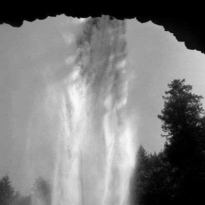Black-and-white photograph, blurred by mist, of a dark waterfall pouring beneath an overhanging rock ledge fringed with conifers.