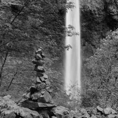Black-and-white photograph of a small rock cairn standing before a thin, streaked waterfall descending a moss-lined cliff in dense forest.