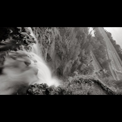 Black-and-white photograph, taken sideways, of a waterfall pouring over a cliff beside a forested slope, a parking area and highway visible below.