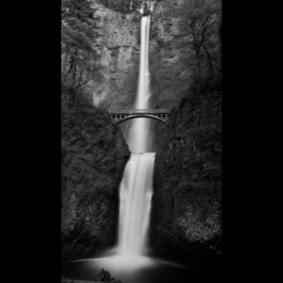 Black-and-white photograph of Multnomah Falls, its two tiers connected by the stone Benson Bridge, plunging into a driftwood-strewn pool.