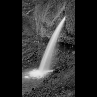 Black-and-white photograph of a waterfall shooting outward from a cave opening in a cliff face, hikers visible walking on the trail far below.