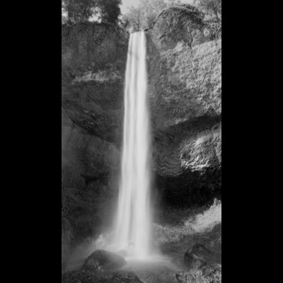 Black-and-white photograph of a tall waterfall dropping past columnar basalt rock formations into a shaded pool below.