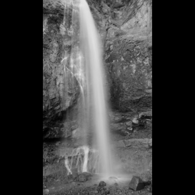 Black-and-white photograph of a narrow waterfall streaming down a sandstone canyon wall into a dark pool at its base.