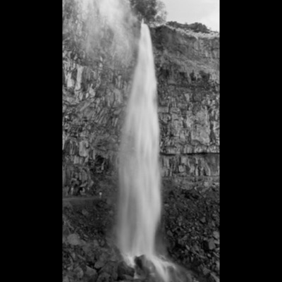 Black-and-white photograph of a tall waterfall pouring off a cliff edge fringed by a single tree, with a tiny figure standing at its base for scale.