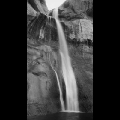 Black-and-white photograph of a narrow desert canyon waterfall streaming down a striated sandstone wall into a dark pool.