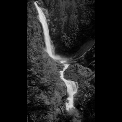 Black-and-white photograph of a mountain waterfall cascading down a rocky slope in stages, framed by dense conifer forest.