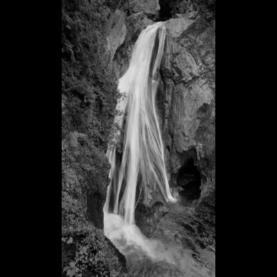 Black-and-white photograph of a wide sheet waterfall pouring off a dark cliff face beside a cave opening.