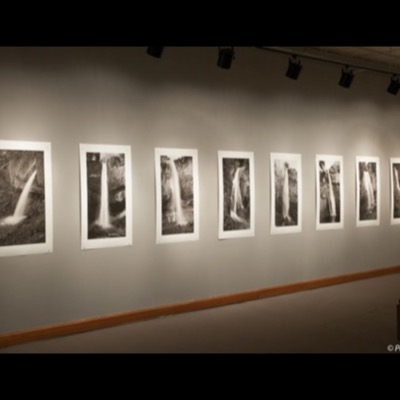 Color photograph of a gallery installation: eight framed black-and-white waterfall photographs from "The Falls" hung in a row along a gray wall under track lighting.