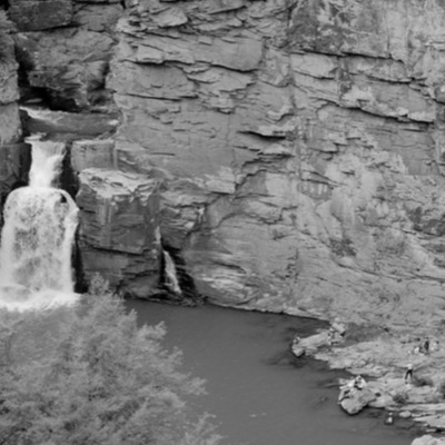 Black-and-white photograph, from above, of a climber ascending a sheer rock wall beside a tall waterfall, other visitors resting on rocks along the pool’s edge below.