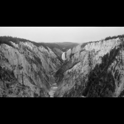 Black-and-white panoramic photograph of the Grand Canyon of the Yellowstone, its waterfall visible far down the canyon between eroded, pale cliff walls.