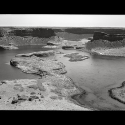 Black-and-white photograph of Dry Falls, a massive ancient cataract in the Columbia Basin, its plunge pools and rock islands spreading across a wide desert canyon floor.
