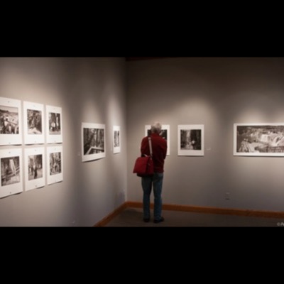 Color photograph of a museum visitor in a red sweater viewing framed black-and-white waterfall photographs, including a contact-sheet grid and a wide panorama, hung across two gallery walls.