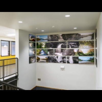 Photograph of "The River" photo-panel installation mounted on a stairwell wall at Nooksack Valley Middle School, four rows of framed river and waterfall images in black-and-white and color wrapping around the corner.