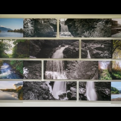 Photograph looking up a corner of the installation from below, showing river and cloud images on the left wall and waterfall and forest panels on the right, framed by the stairwell ceiling and railing.